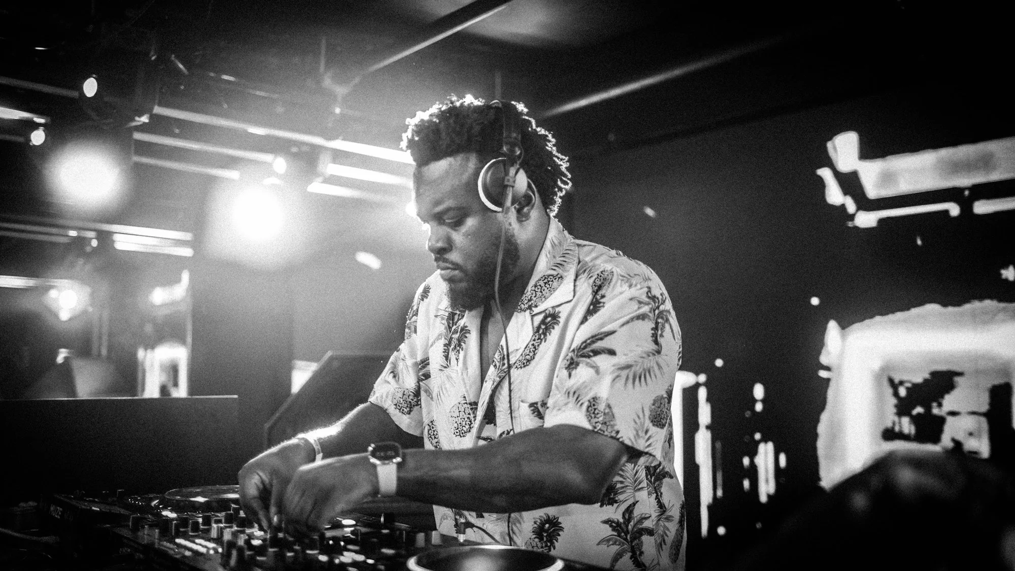 A DJ with headphones, wearing a Hawaiian shirt, is working at a DJ booth in a nightclub with bright lights and a dark background.