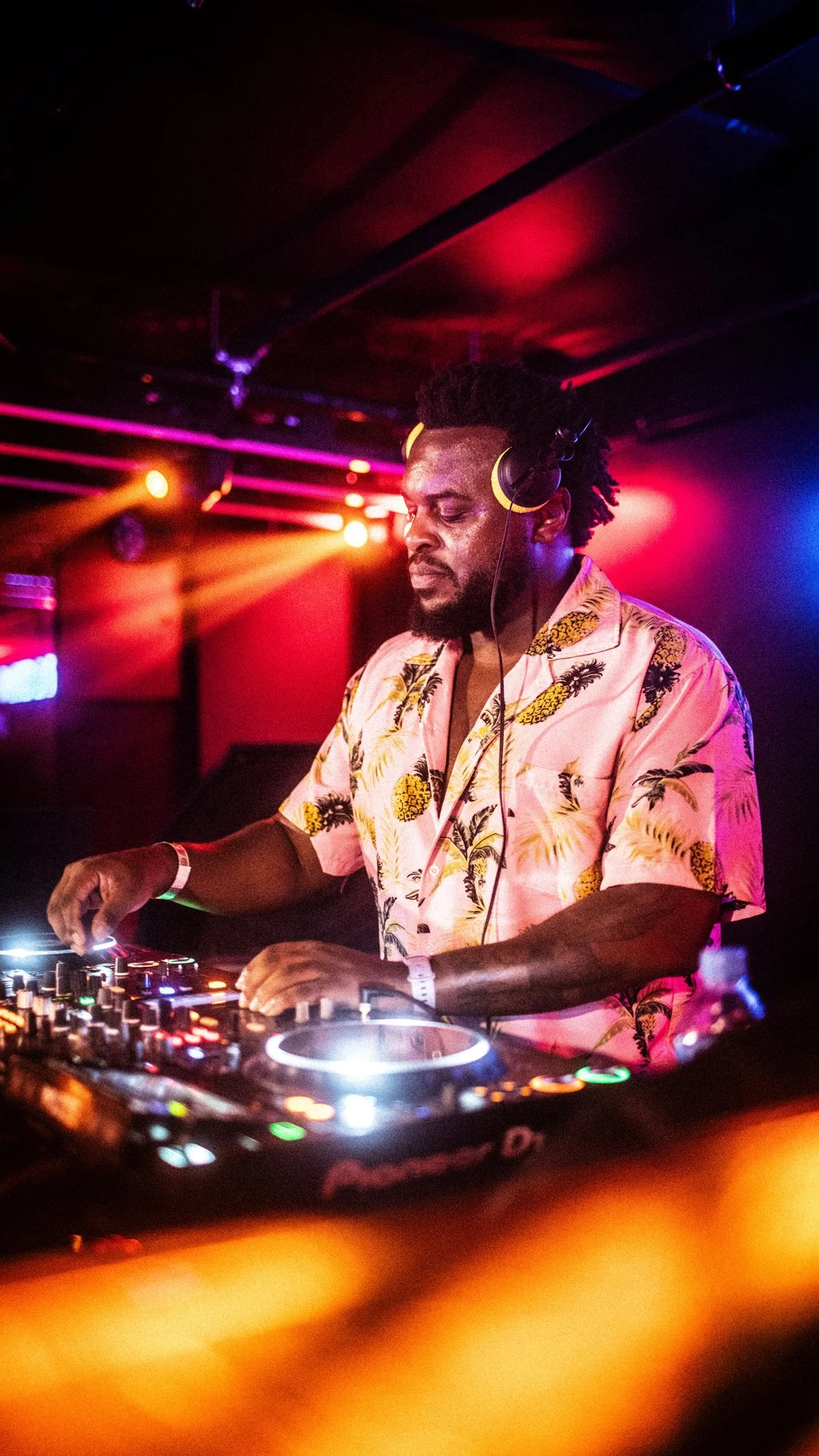 A male DJ wearing a pink Hawaiian shirt with pineapples, headphones, and a white wristband, performing behind a DJ console with colorful lights and a dark, vibrant club background.