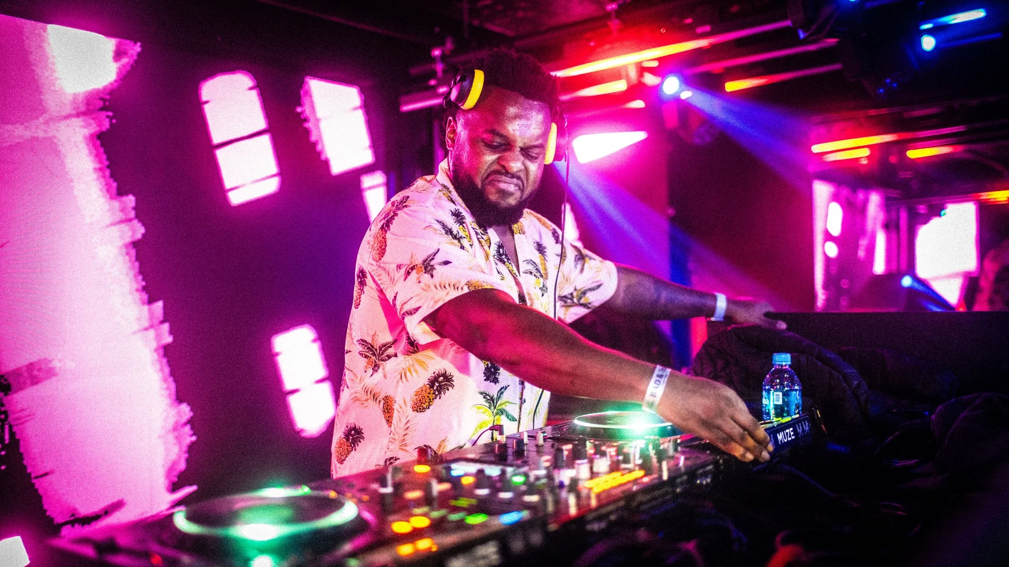 A DJ wearing a tropical shirt and yellow headphones playing music at a vibrant nightclub with colorful lights and a digital backdrop.