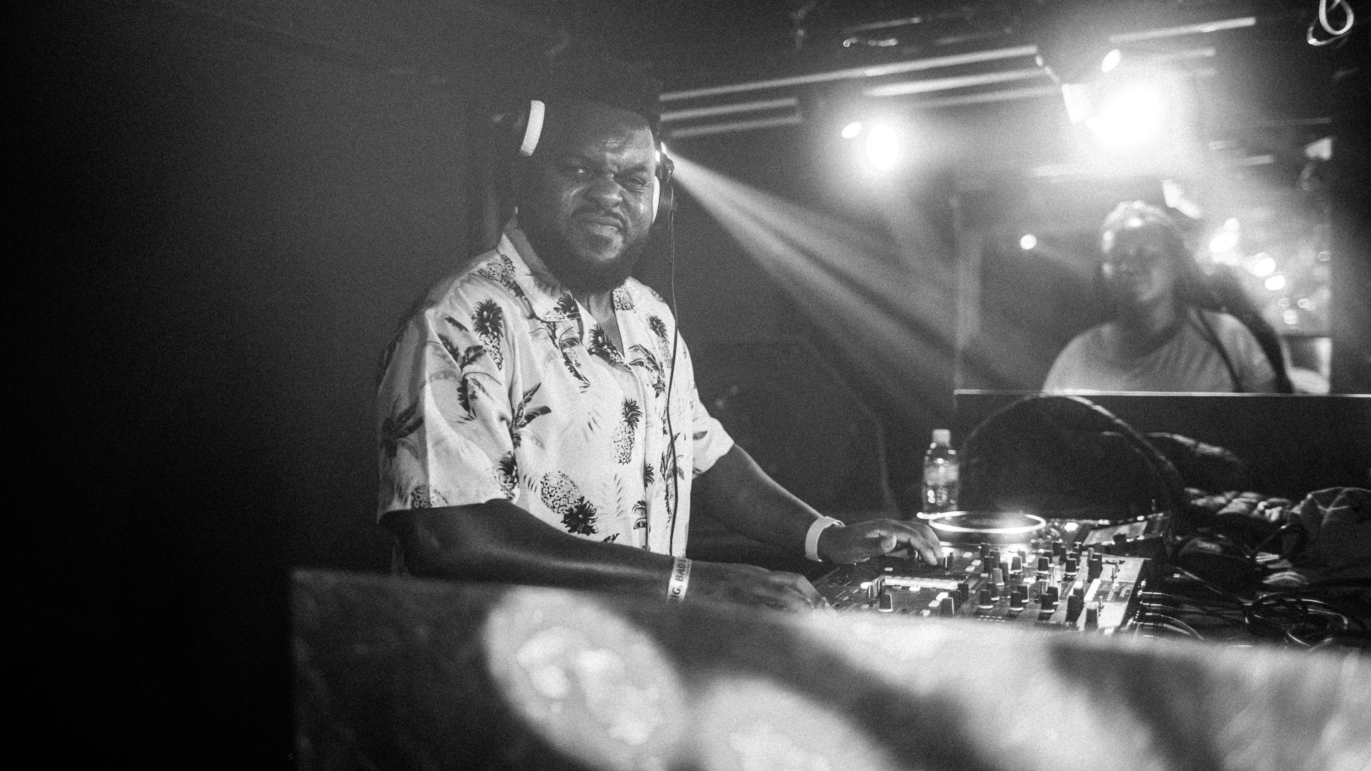 A DJ wearing a Hawaiian shirt and headphones stands at a mixing console in a nightclub, with another person smiling in the background, illuminated by stage lights.