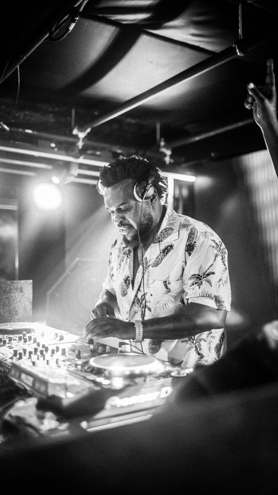 A DJ with headphones, wearing a Hawaiian shirt, performing at a nightclub or music event in black and white.