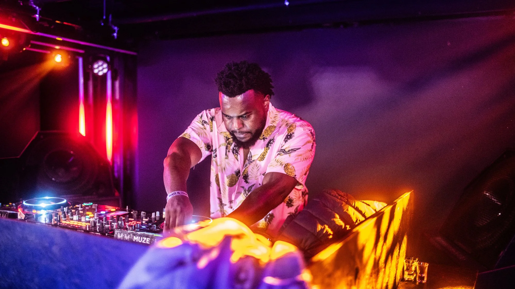 A DJ in a tropical shirt performing at a nightclub with vibrant red, purple, and orange lighting.