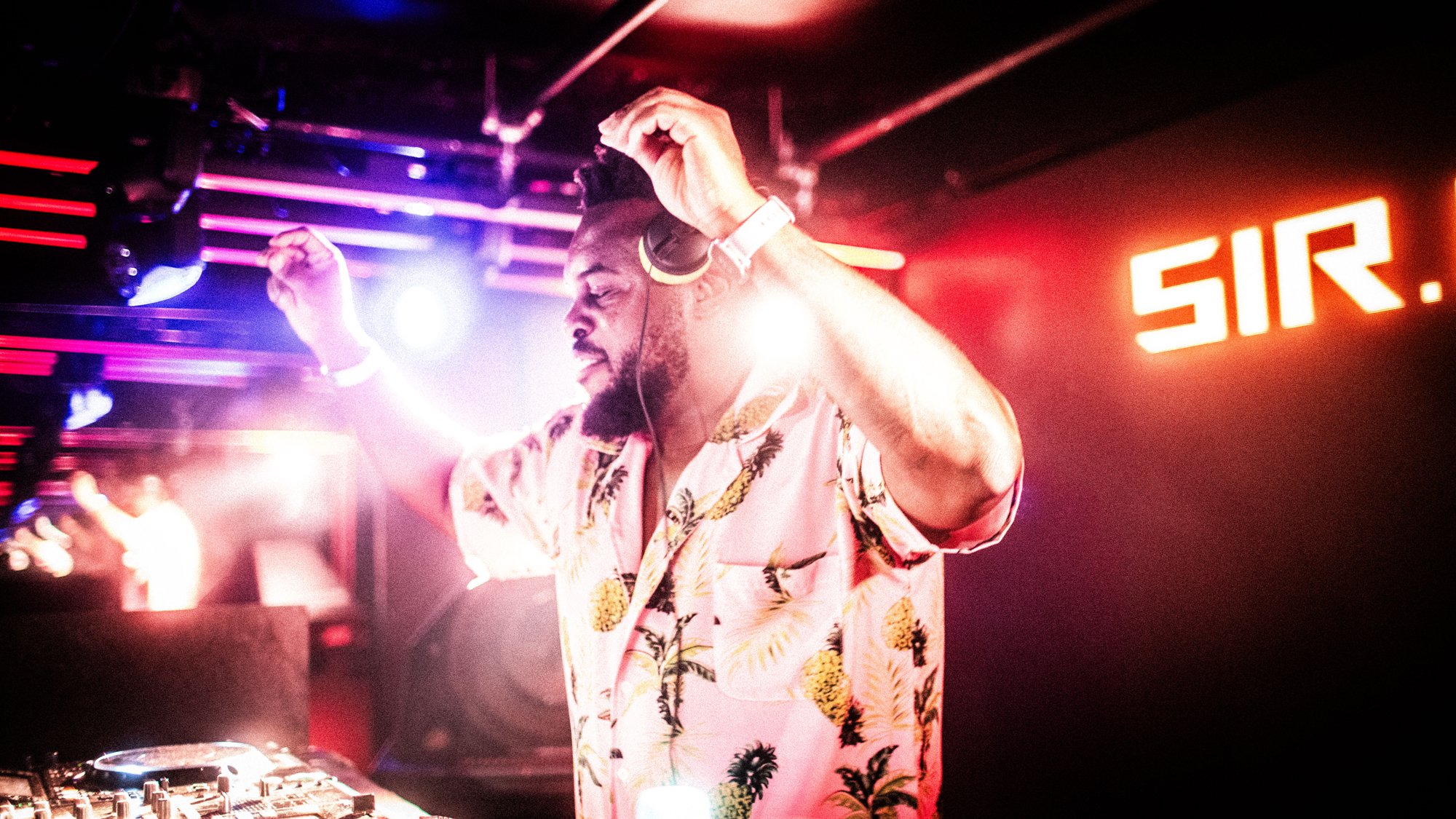 DJ wearing tropical print shirt, headphones, with arms raised, performing at nightclub with colorful lights and Sir sign in background.