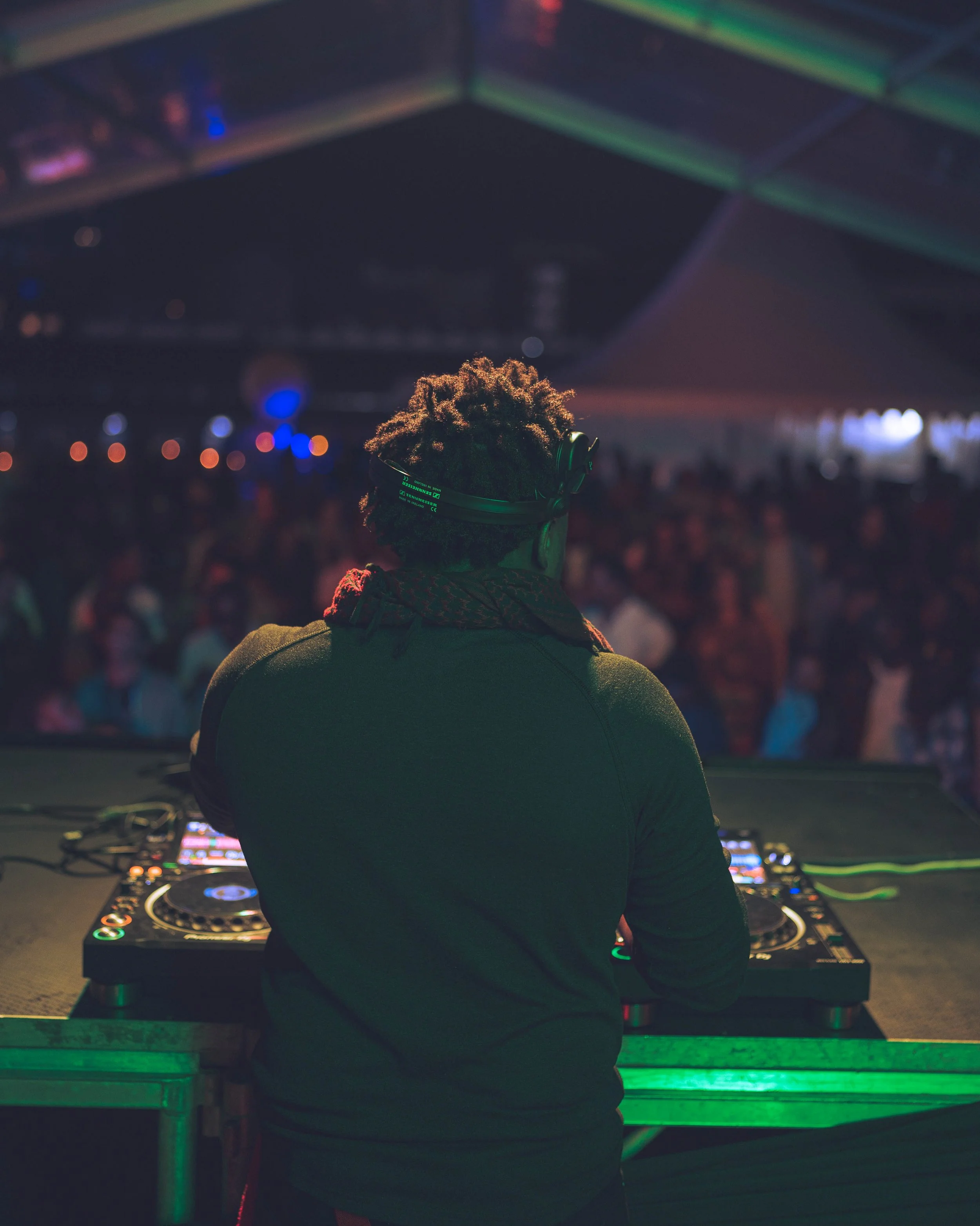 A DJ wearing headphones is performing at a nightclub with a crowd dancing in the background.