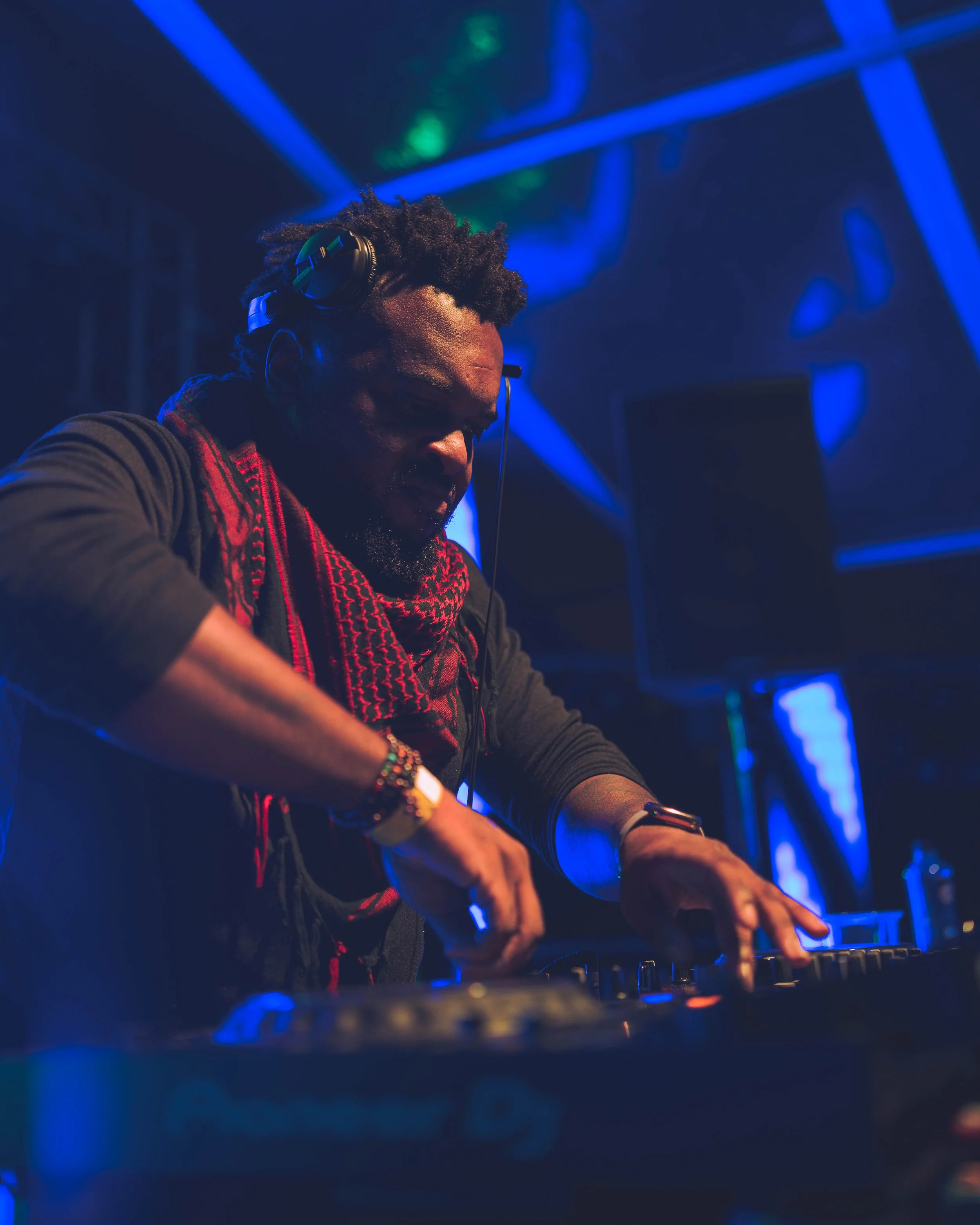 A DJ wearing headphones and a red scarf performs with a mixing console in a dimly lit club with blue and green stage lights.