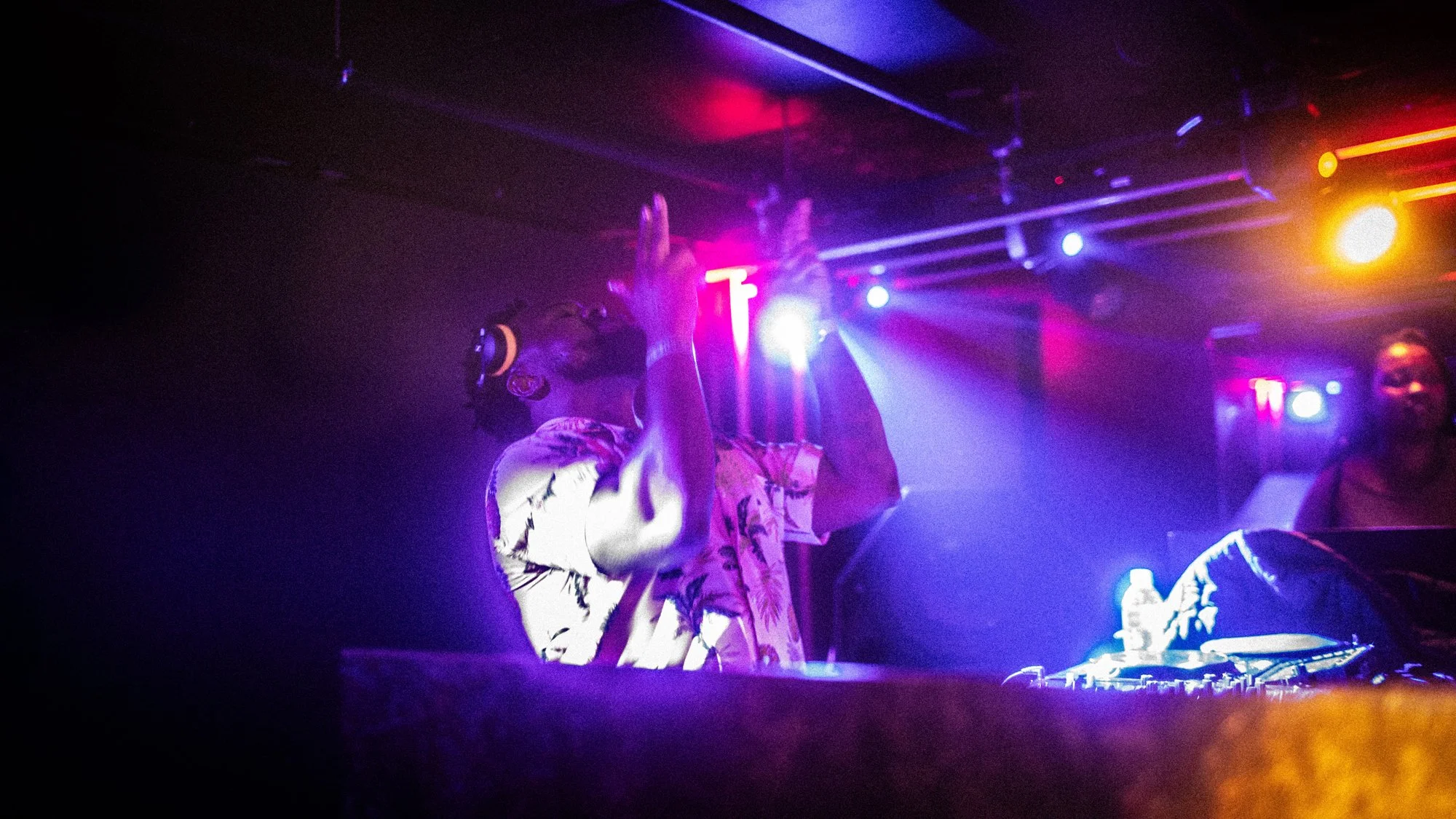 A DJ wearing headphones and a tropical shirt is performing, with one hand raised, in a club with colorful lights and people dancing.