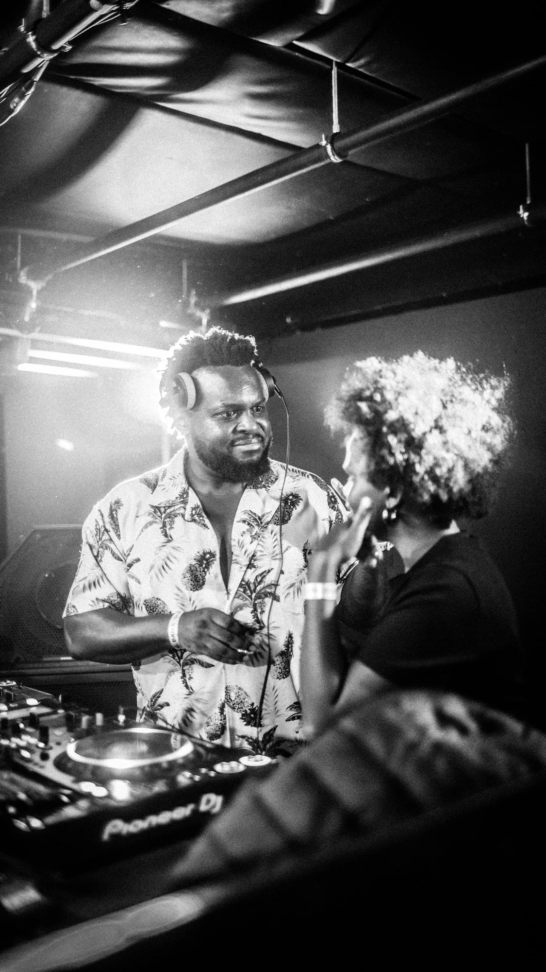 Black and white photo of a DJ wearing headphones and a patterned shirt, standing behind DJ equipment, talking to a woman with curly hair at a nightclub or music event.