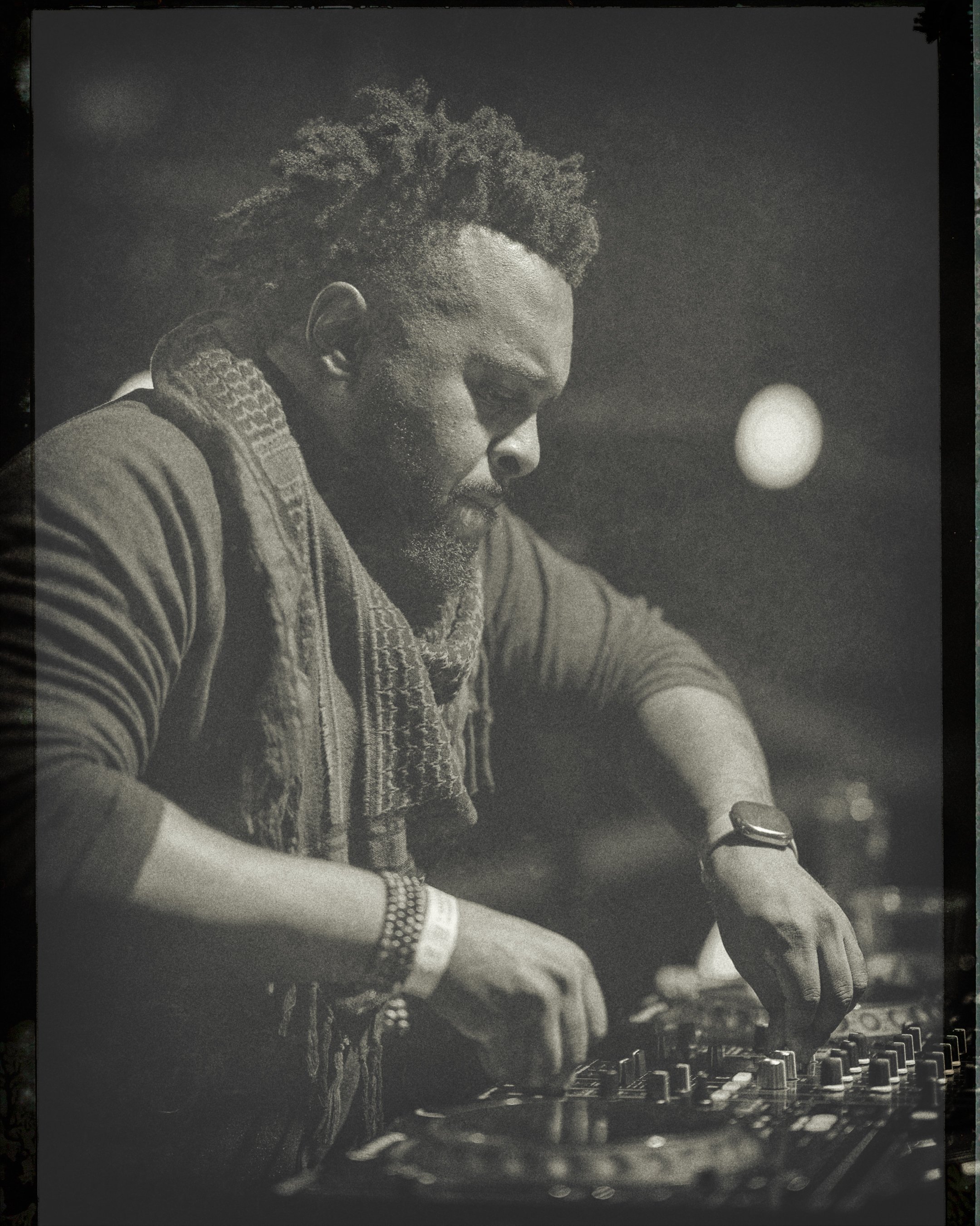 A man with a beard, wearing a wristwatch and bracelets, is DJing at a music event, focused on mixing music on a DJ console with a blurred background and a round light