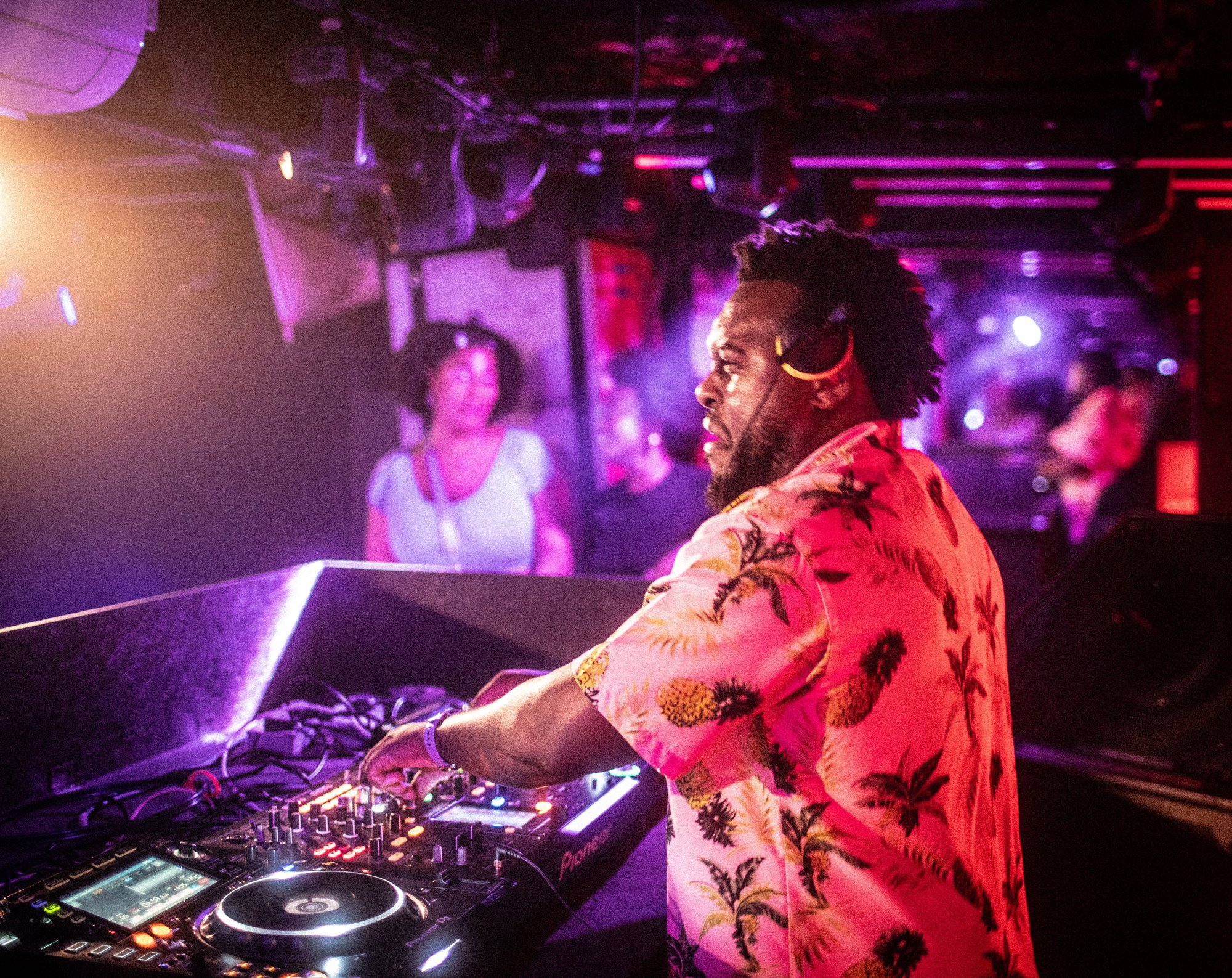 A DJ with a beard and dreadlocks wearing headphones around his neck, operating a DJ mixer at a nightclub with purple and pink lighting. There are people dancing and enjoying the music in the background.