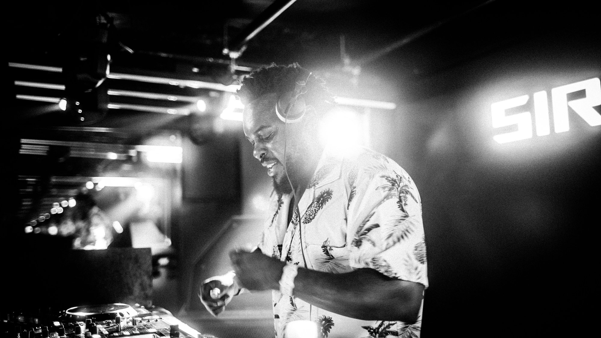 A black and white photo of a DJ wearing headphones, smiling, and mixing music at a club or party. The background has blurred lights and a sign that reads 'SIR'.