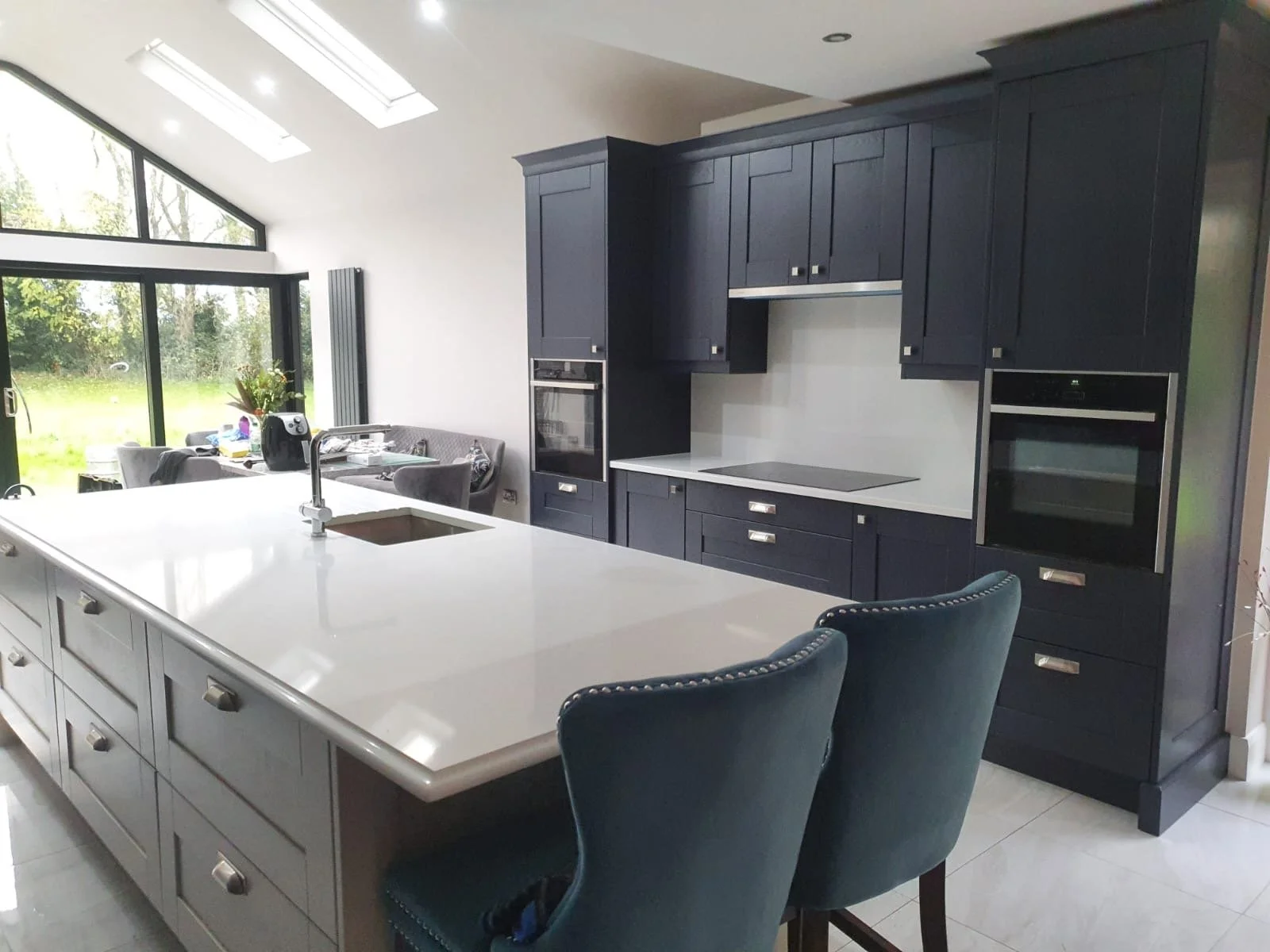 Modern kitchen with white island counter, dark blue cabinets, built-in oven, and a view of a bright dining area with large windows and a sliding door.