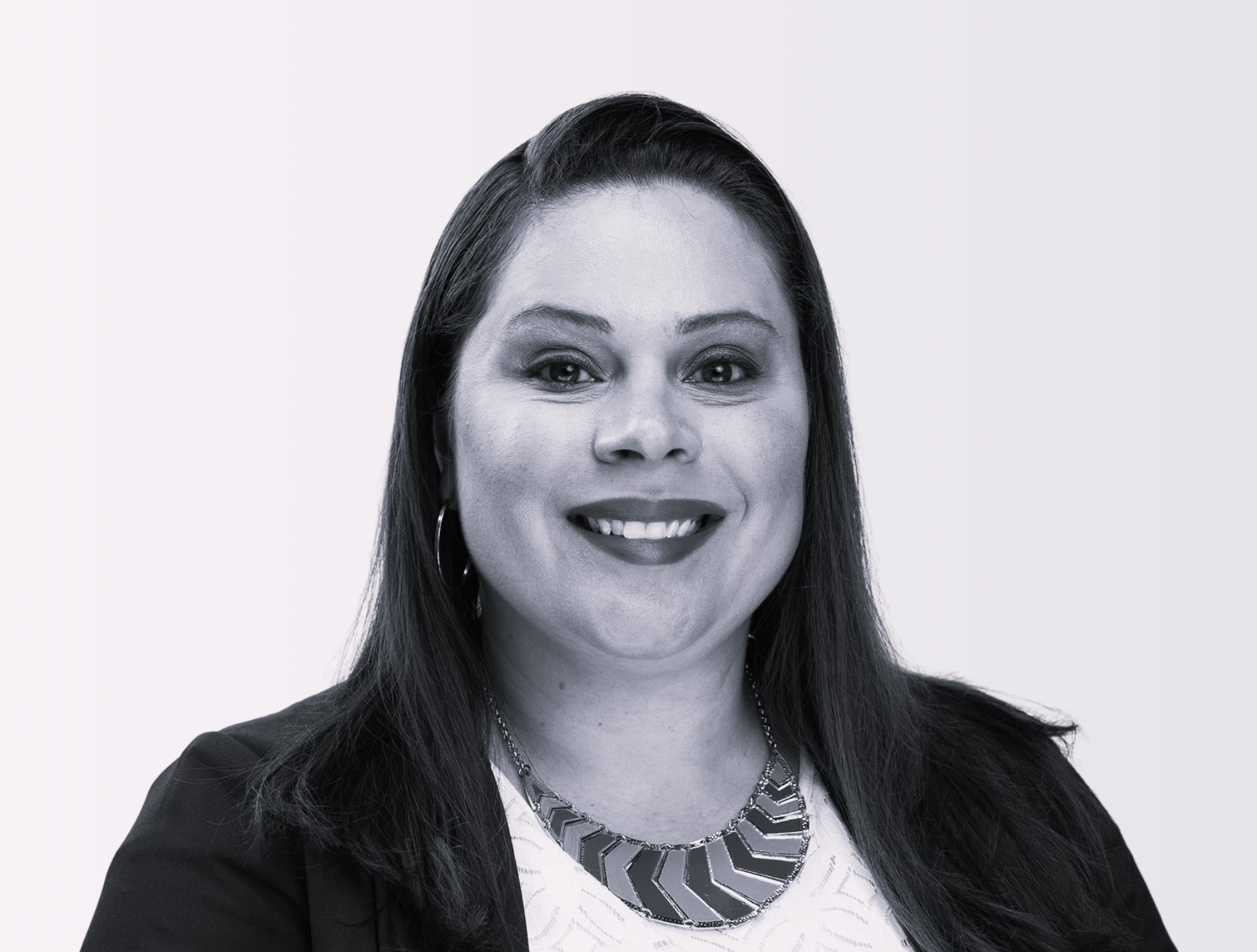 A smiling woman with long dark hair wearing a black blazer, a white patterned top, hoop earrings, and a statement necklace, against a plain light background.