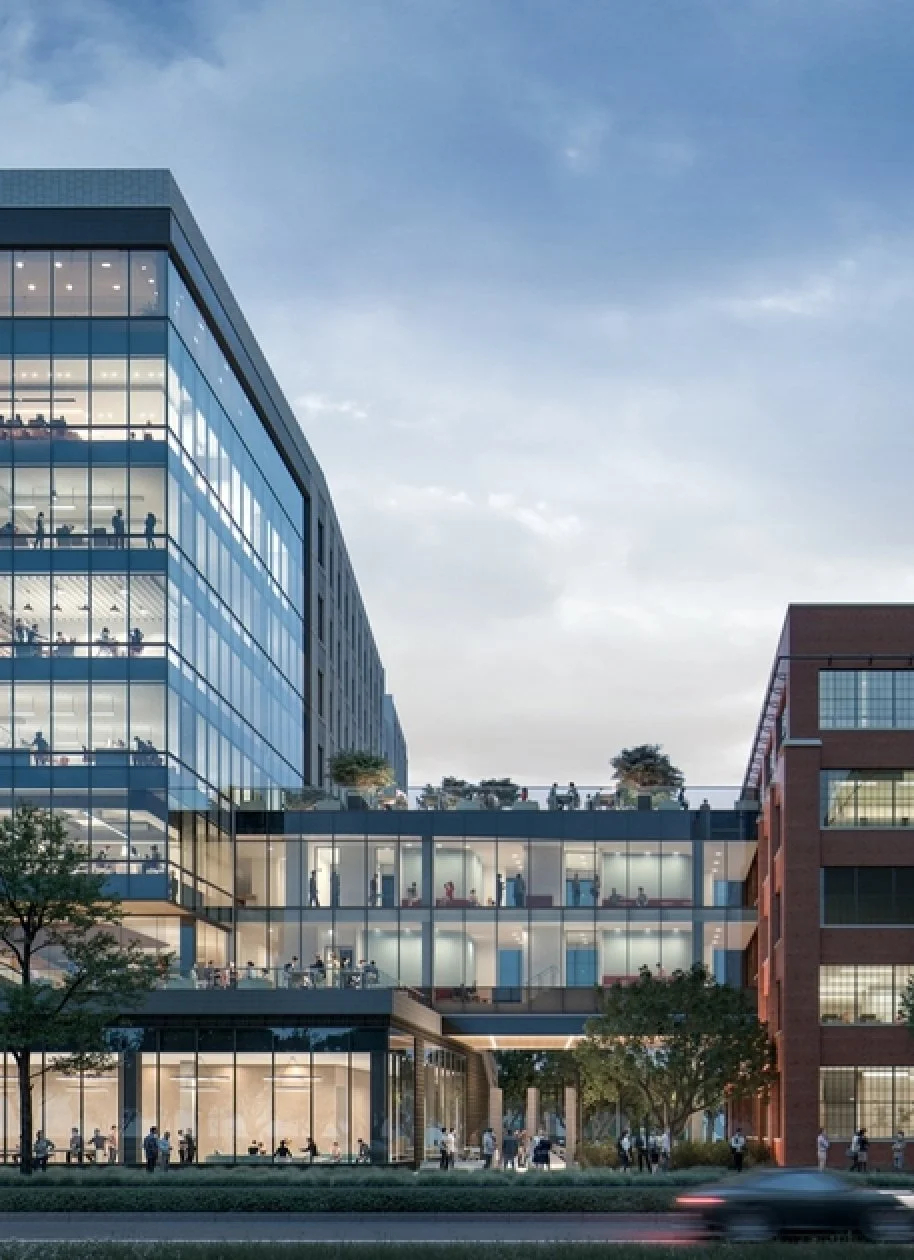 Modern multi-story office building with glass windows, nighttime view showing illuminated offices and people working inside, and trees in the foreground.
