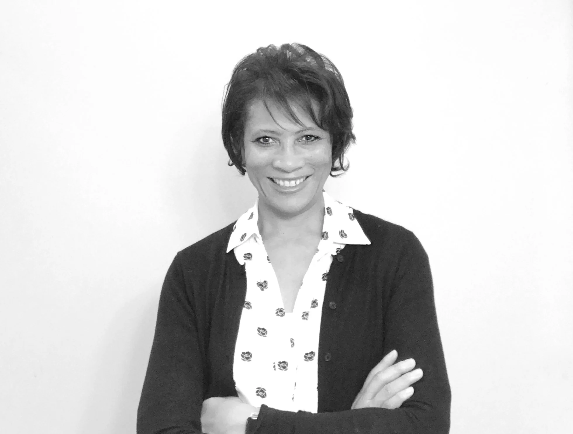 A woman with short hair smiling and standing with arms crossed, wearing a patterned blouse and a dark cardigan, against a plain white background.