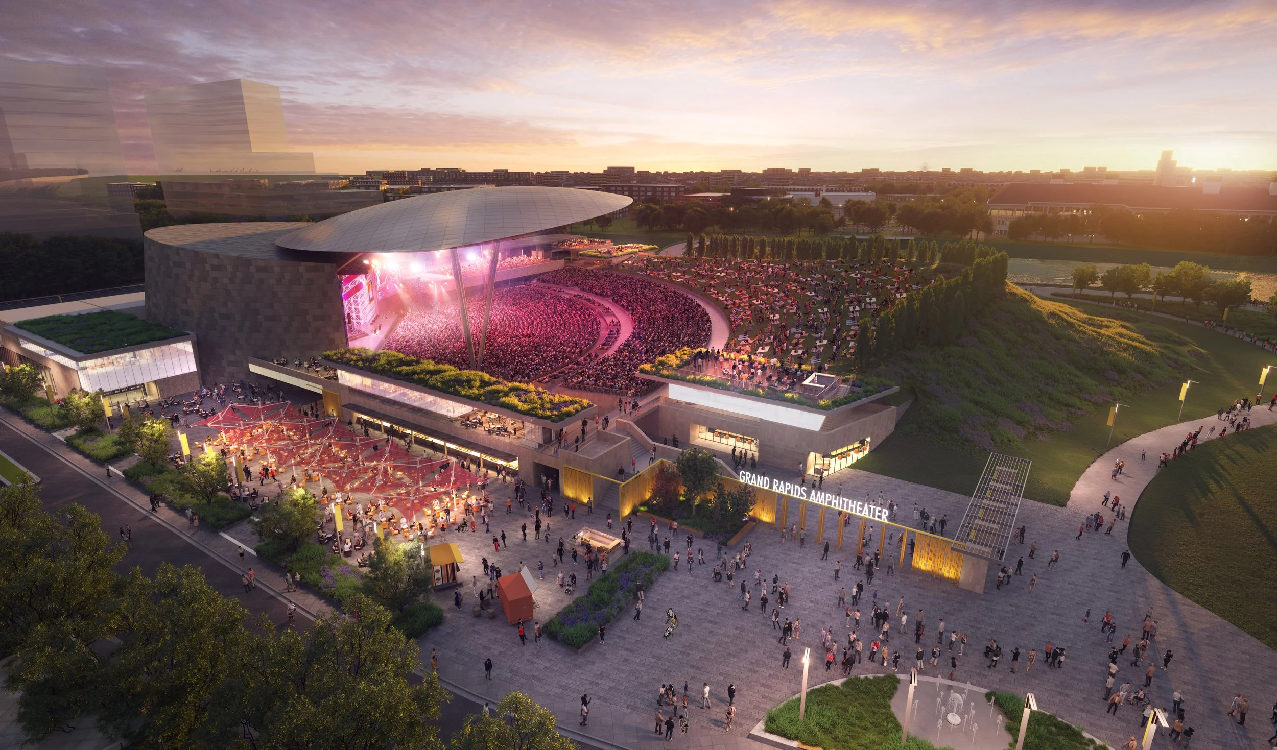 An aerial view of the Grand Rapids Amphitheater at sunset, with a large crowd attending a concert on the open-air stage, surrounded by trees, pathways, and green hills.