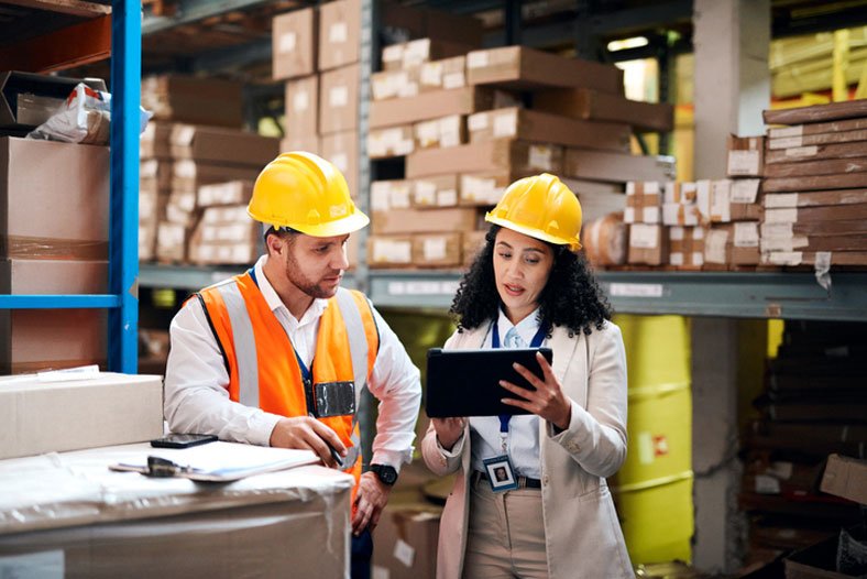 Two warehouse workers, a man and a woman, wearing yellow safety helmets, are discussing something on a clipboard tablet in a storage area with shelves filled with boxes and wooden planks.