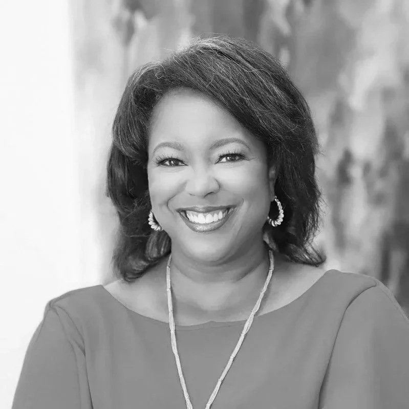 A smiling African American woman with shoulder-length hair, wearing a necklace and earrings, dressed in a top with a rounded neckline, with a blurred background.