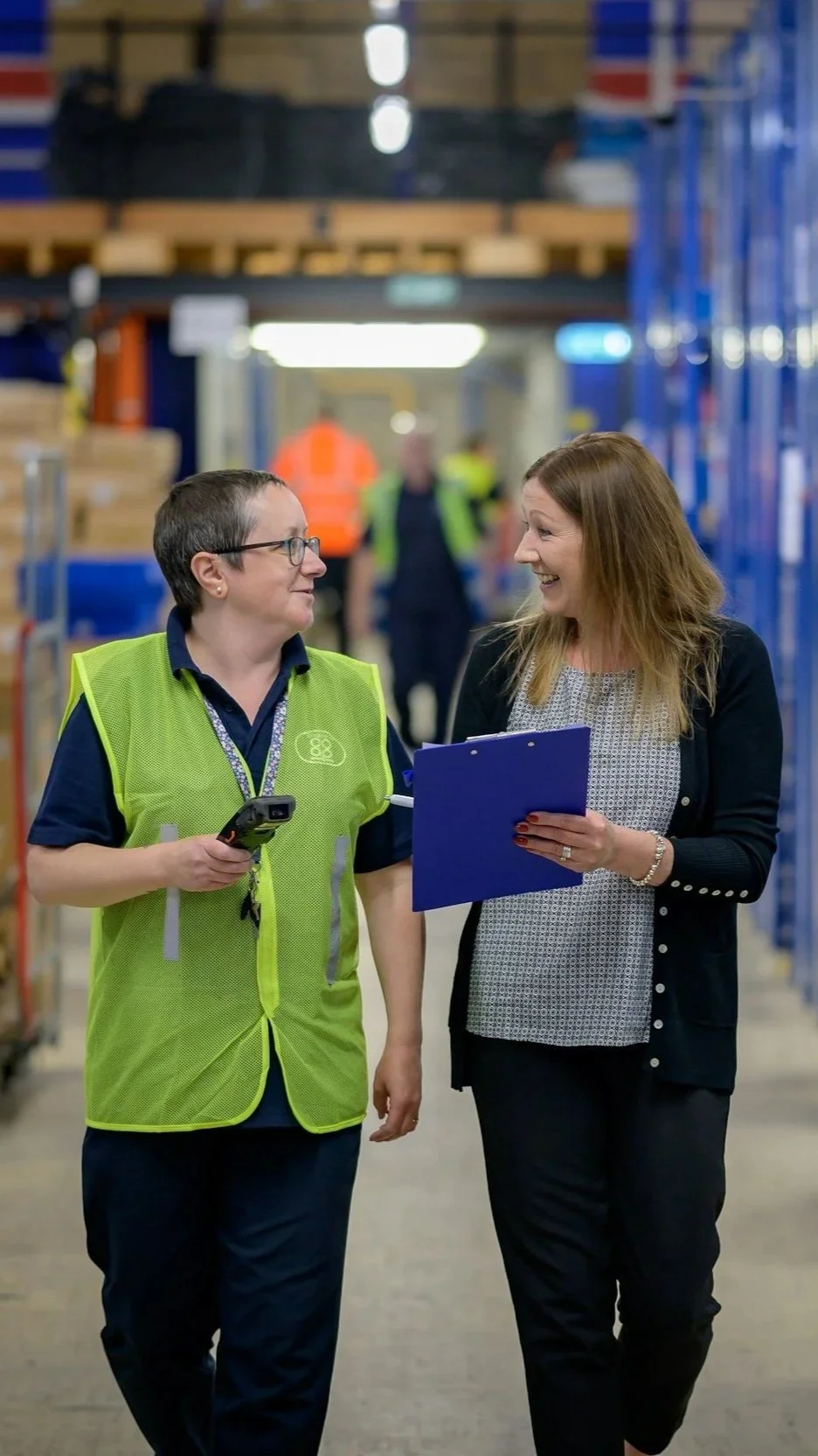 Two women, one wearing a green safety vest and the other holding a blue clipboard, are walking and talking in a warehouse aisle with shelves filled with boxes and workers in the background.