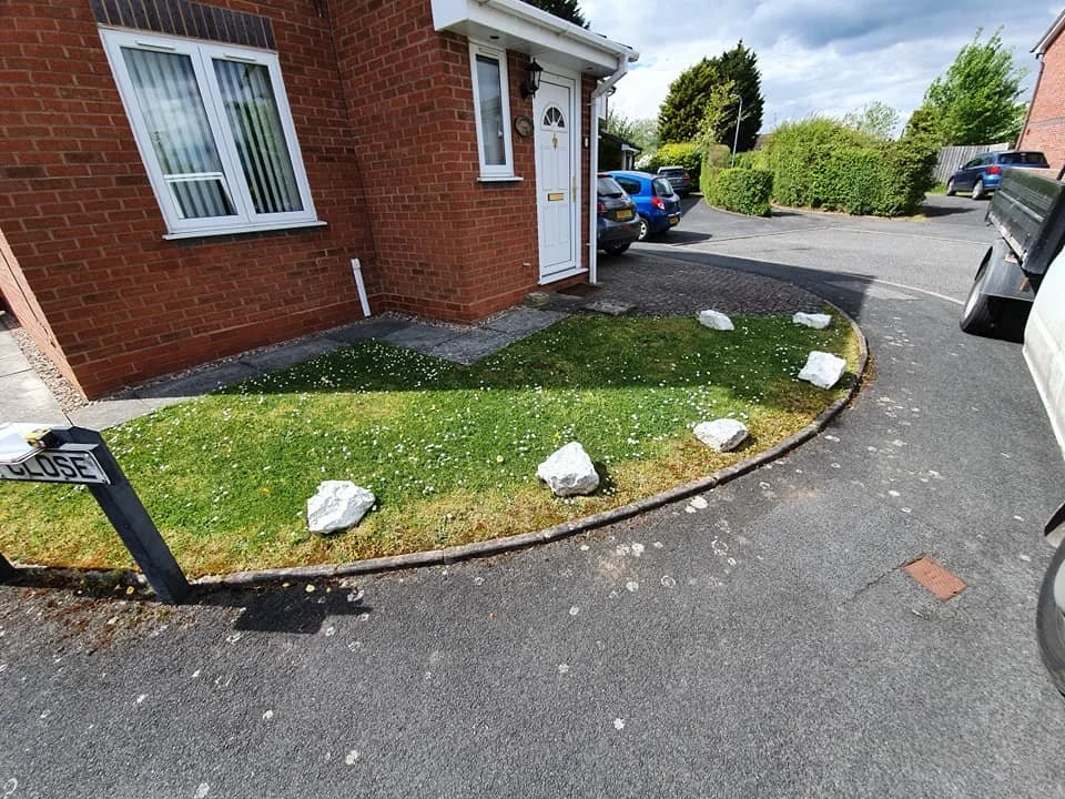 A small grassy yard with white rocks along the edge, outside a brick house, next to a driveway with parked cars.