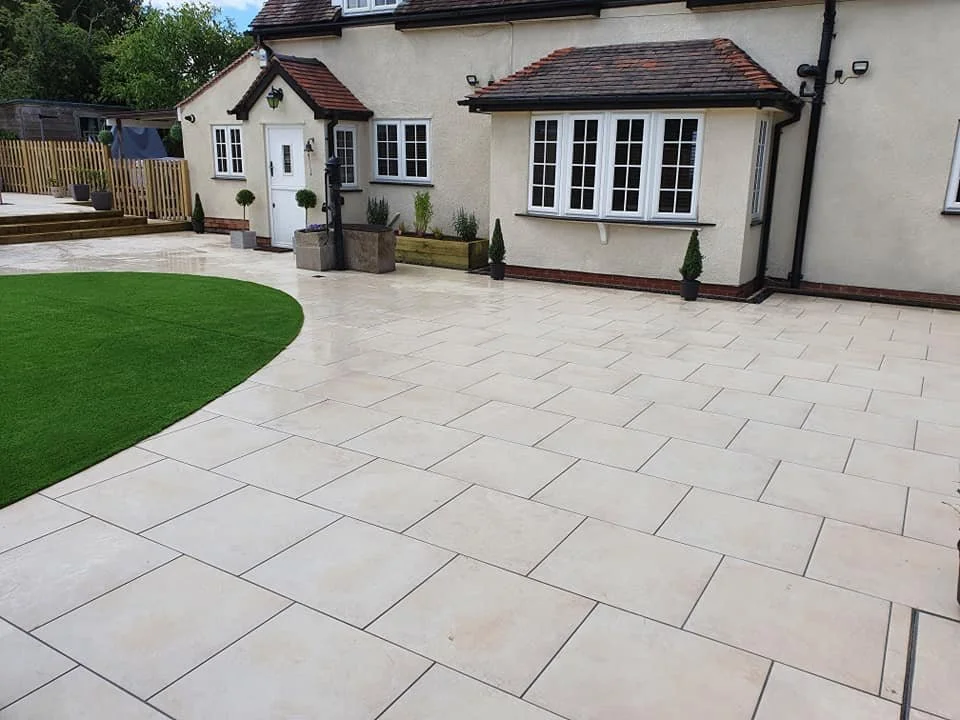 Backyard patio with beige paving stones, green grass patch, potted plants, and a white house with a bay window.