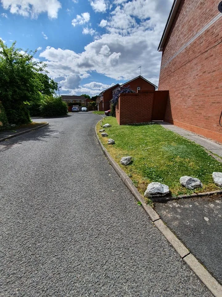 A curved asphalt road running past brick residential buildings with a small grass area and large rocks lining the edge, under a partly cloudy sky.