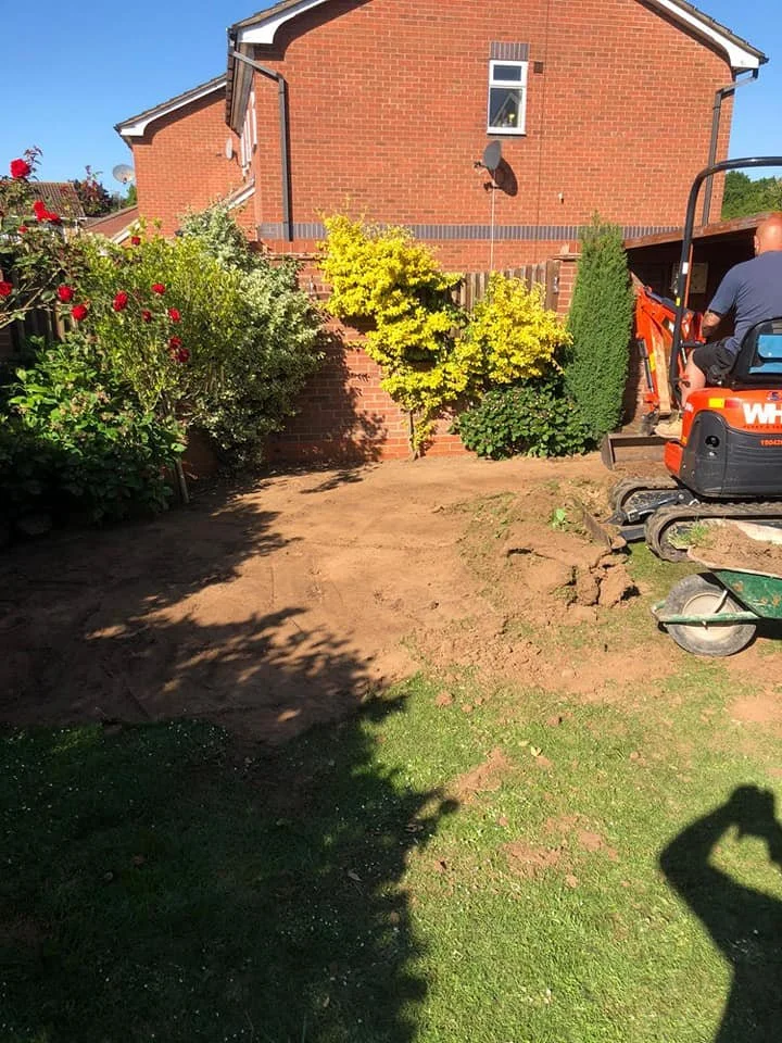 A backyard garden undergoing landscaping, with a small excavator and a person operating it, surrounded by plants, shrubs, and a brick house in the background.