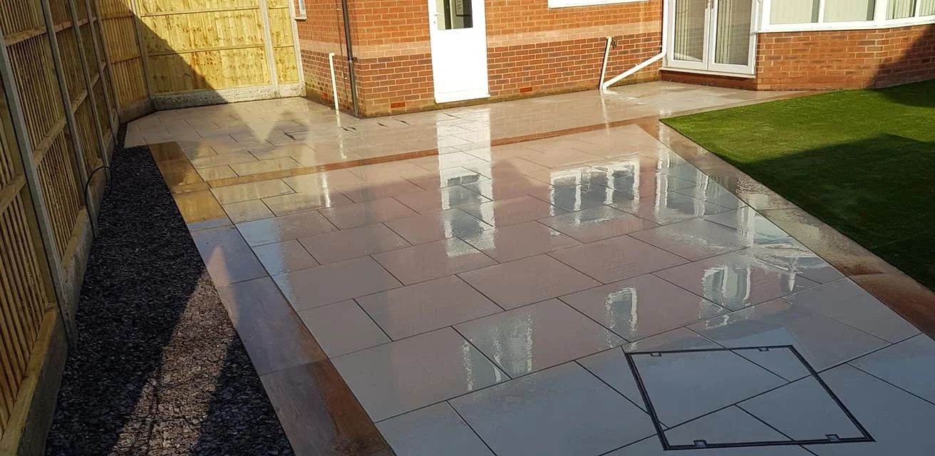 Recently cleaned and wet outdoor patio area with large rectangular tiles, near a brick house with sliding glass door, small patch of green grass, wooden fence on one side, and drainage hatch in tile surface.