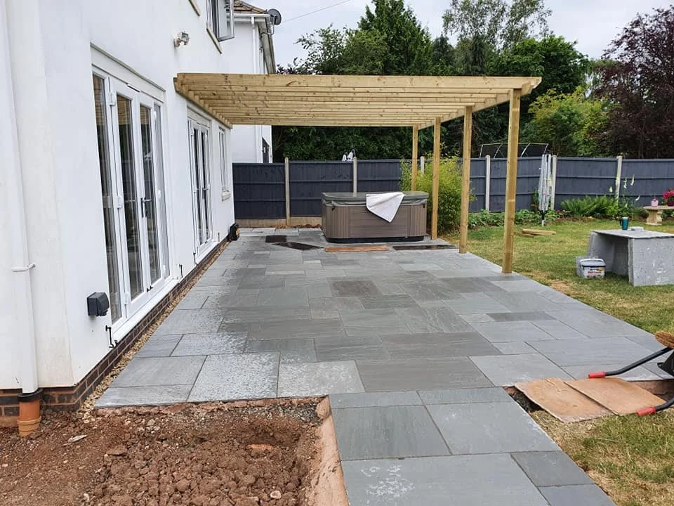 Backyard patio under construction with newly laid gray stone pavers, a hot tub under a wooden pergola, and a grassy area with a garden and fence in the background.