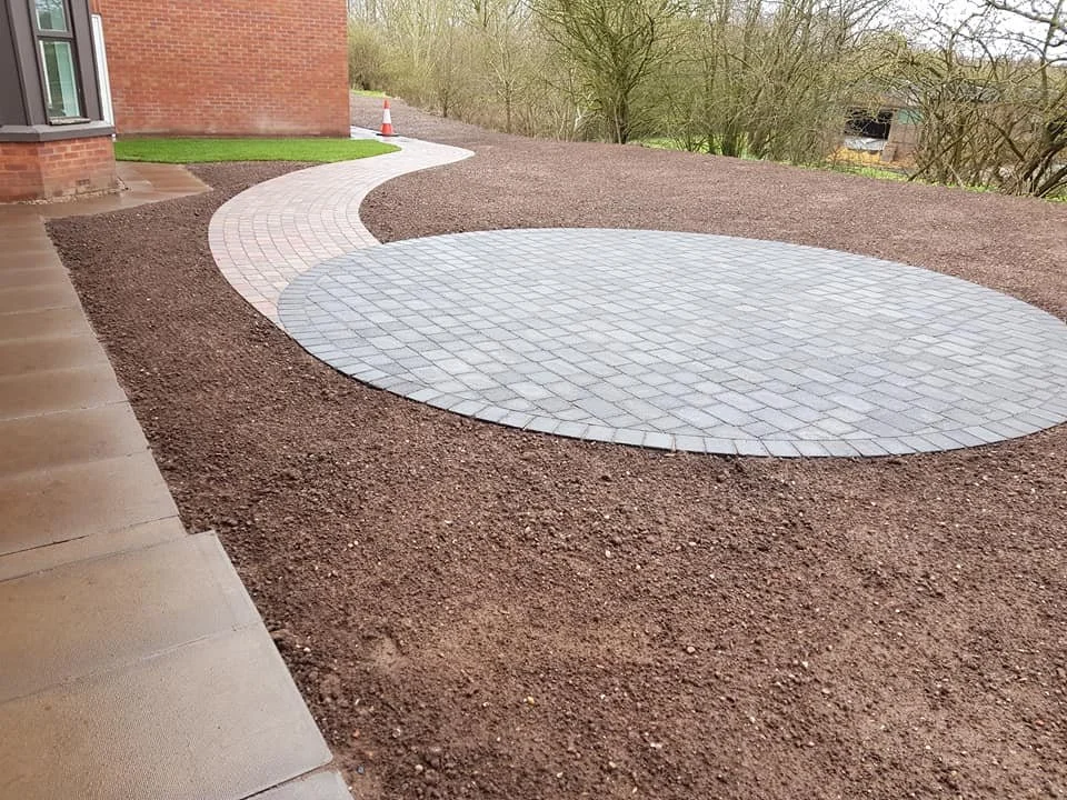 Curved concrete and brick pathway with a flower bed on the side, leading away from a brick building in a yard under construction.