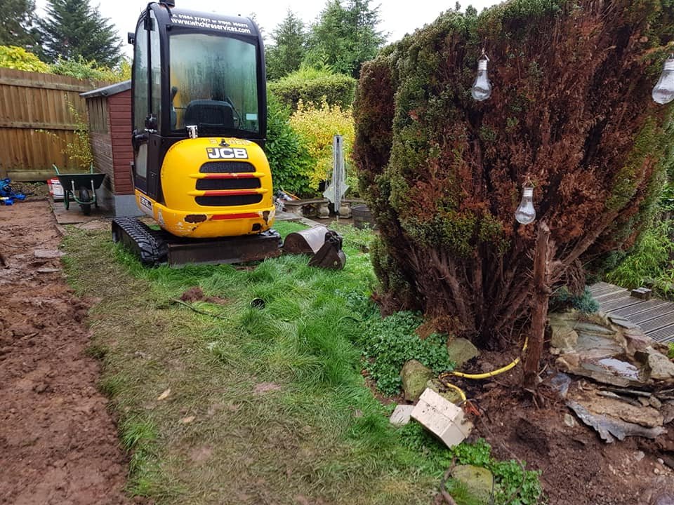 A small yellow JCB excavator parked on a grassy area in a backyard, with a large bush and decorative lights on it, and various gardening tools and materials around.