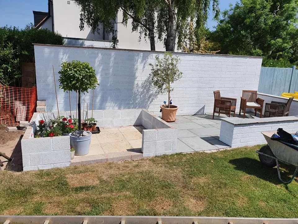 A small backyard garden area with a white brick wall, potted plants including a flowering plant and a tree, outdoor seating with wooden chairs, a wheelbarrow on the grass, and some construction materials.