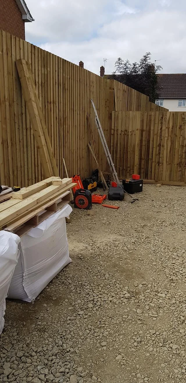 A backyard with a new wooden fence under construction, various tools and supplies including a ladder, saw, and wood planks are scattered on the ground and leaning against the fence, with a house and trees in the background.