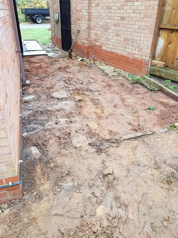 A backyard area with dirt and soil, next to a brick house and a wooden fence, with a black gate and a small patch of grass in the background.