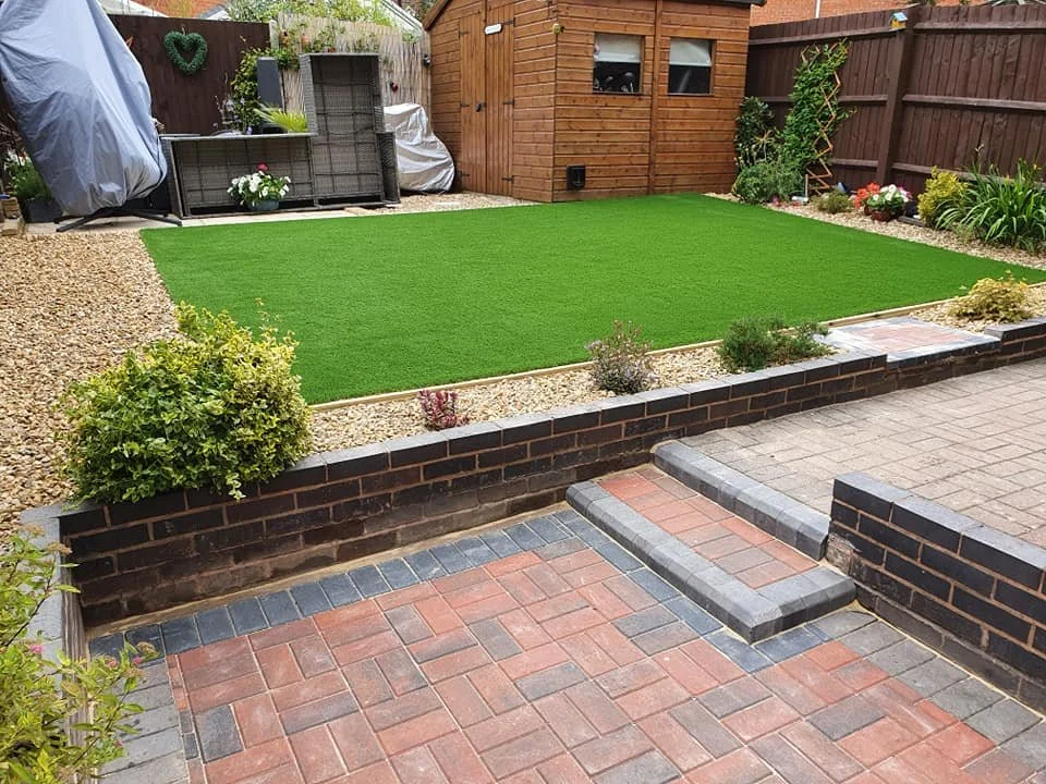 Backyard with a small artificial grass lawn, bordered by gravel and small plants, with a brick and paved patio area, and a wooden shed and garden furniture in the background.