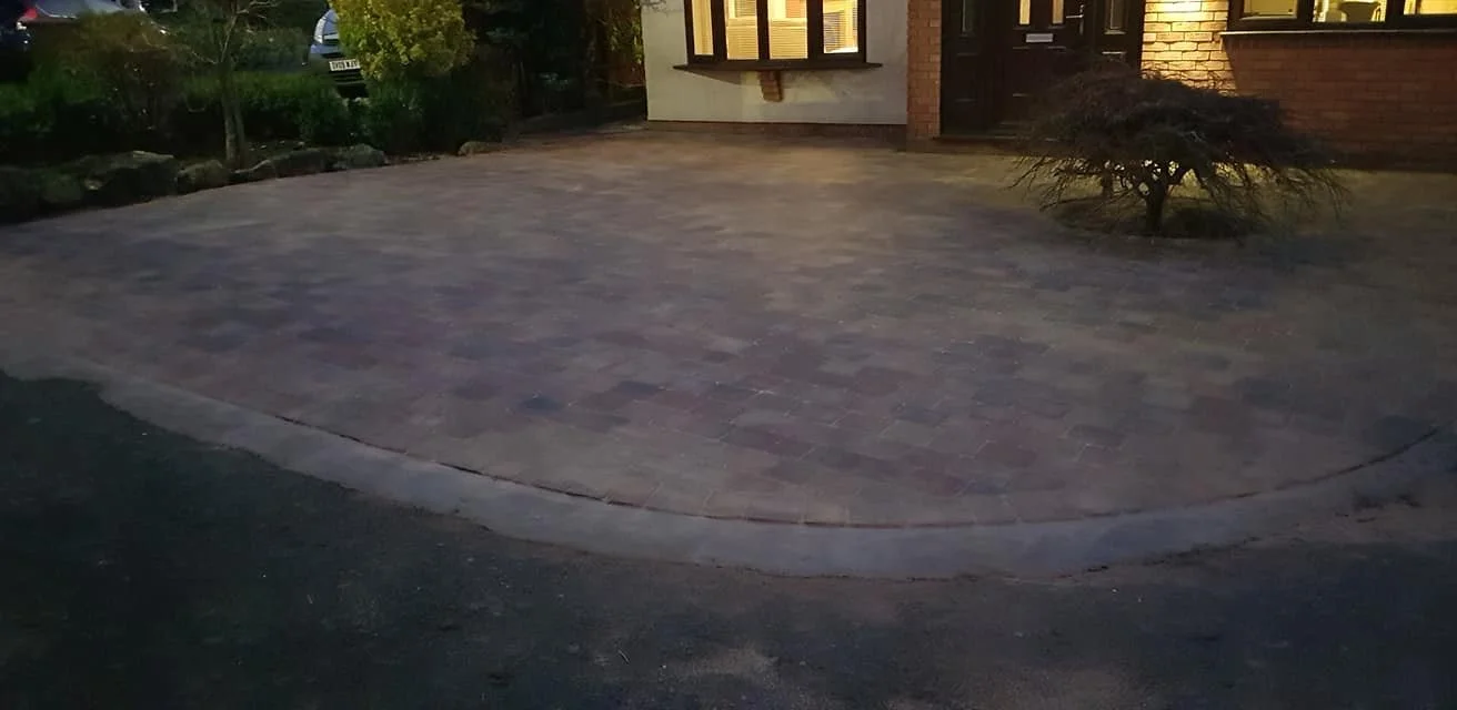 A paved driveway in front of a house with a brick exterior and lit windows, decorated with a small bush on the right and greenery along the left side.