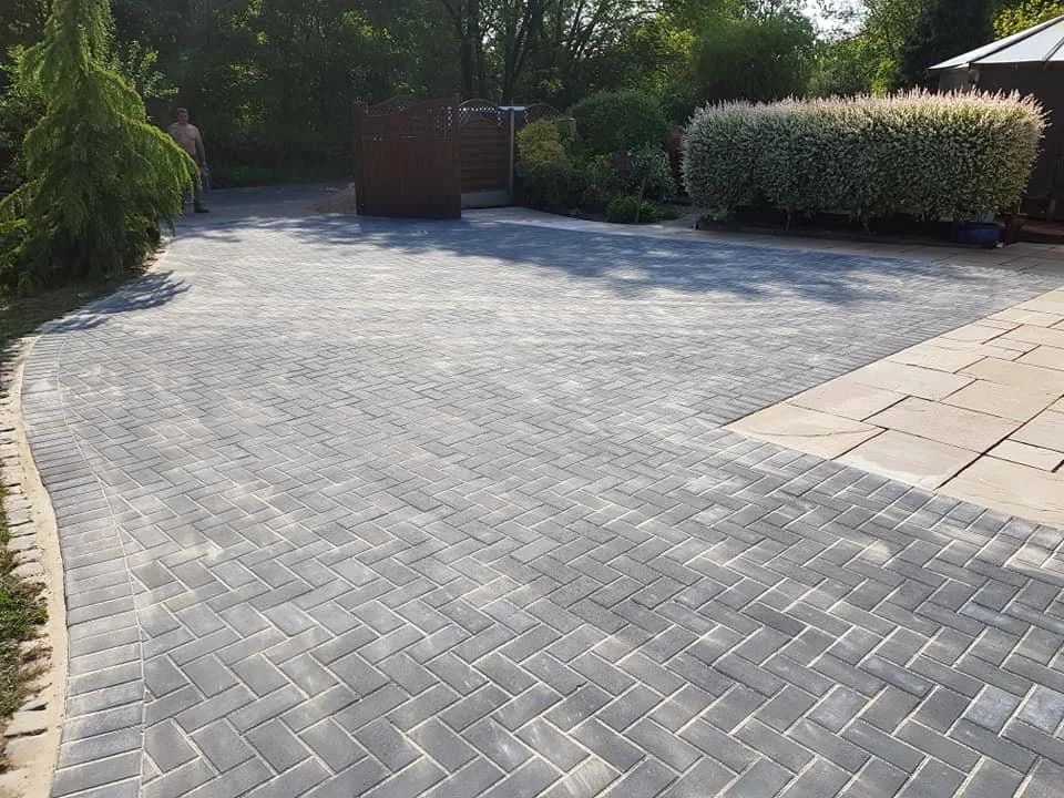 A spacious backyard with a paved driveway made of gray bricks, a wooden gate, and lush green bushes and trees surrounding the area.