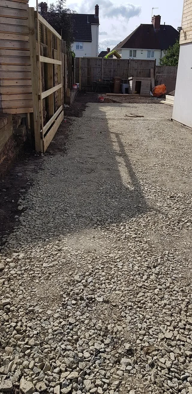 A backyard area under construction with a gravel ground and a wooden fence. There are garden tools, buckets, and a dog house in the background. The sky is partly cloudy.