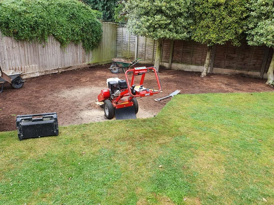 Backyard with a garden bed under construction, a red tiller tool, a wheelbarrow, a shovel, a black toolbox, grass in the foreground, and a wooden fence with trees in the background.
