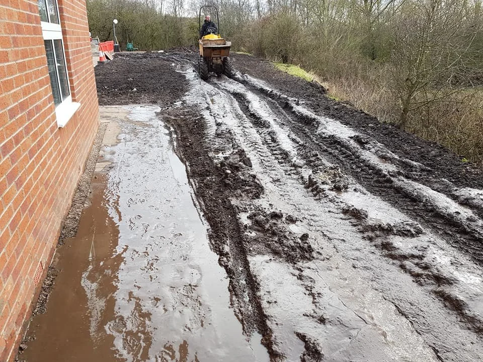 A construction worker operating a small bulldozer, leveling muddy ground outside a brick building.