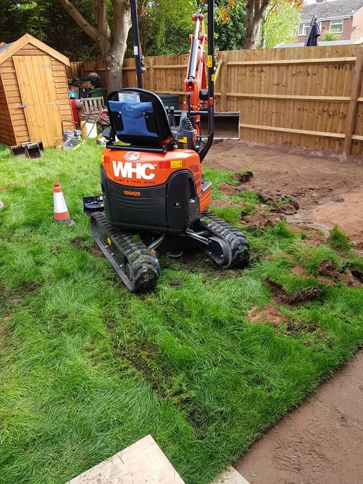 Small orange and black mini excavator on a grassy backyard, digging trenches along a fence line, with a wooden shed and trees in the background.