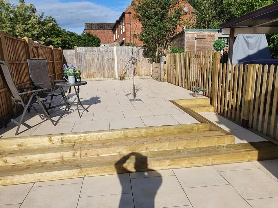 A backyard patio with wooden steps leading up to a paved area, surrounded by a wooden fence. There are two chairs and a small table with potted plants, and a young, leafless tree in the middle.