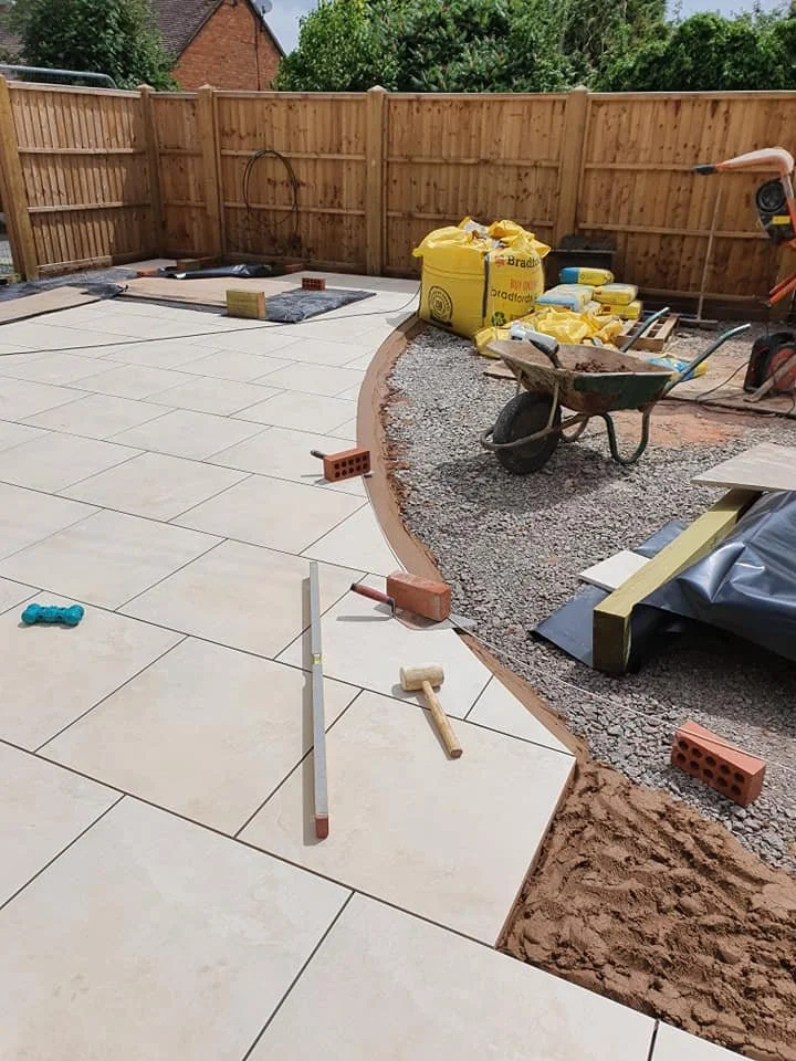 A backyard patio area under construction with beige tiles being laid, construction tools and materials like bricks, a wheelbarrow, and a chalk line visible, and a wooden fence surrounding the workspace.