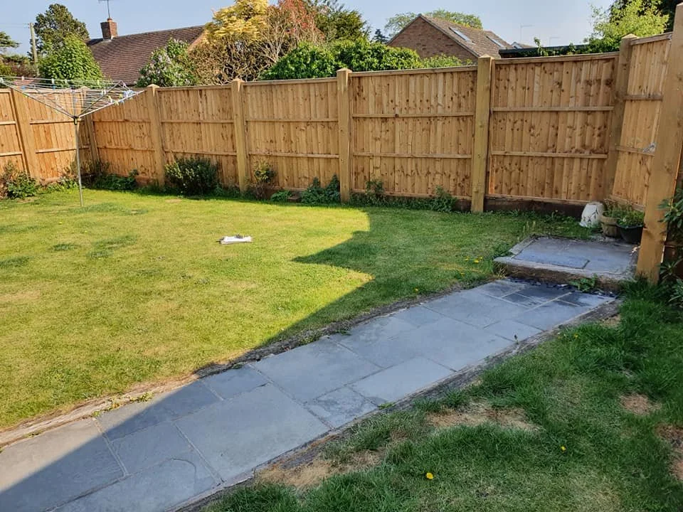 A backyard with a grassy lawn, surrounded by a wooden fence. There is a rotary clothesline in the left background and a partially paved stone pathway running through the yard. Small plants and pots are visible near the fence on the right side.