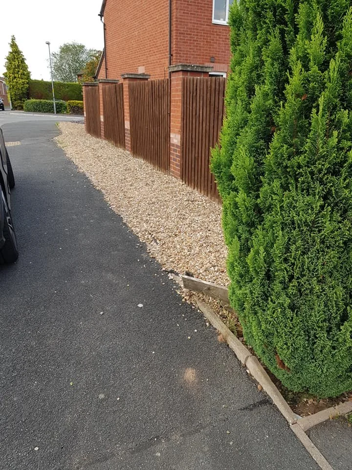 A sidewalk alongside a gravel area, with a brick house on the left, a wooden fence, and a large green bush on the right.