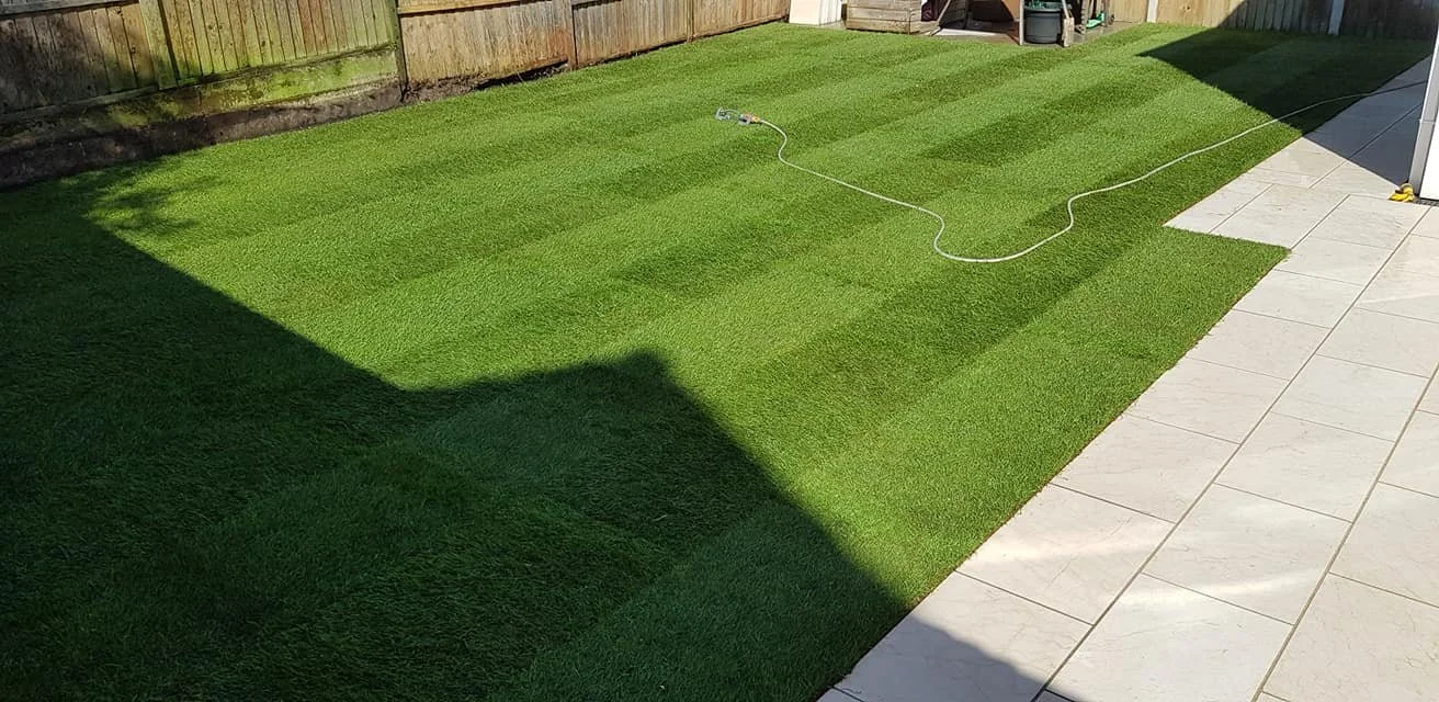 An outdoor backyard scene showing a freshly laid green grass lawn with an alternating striped pattern next to a tiled patio. There is a garden hose lying on the grass, and a wooden fence encloses the yard.