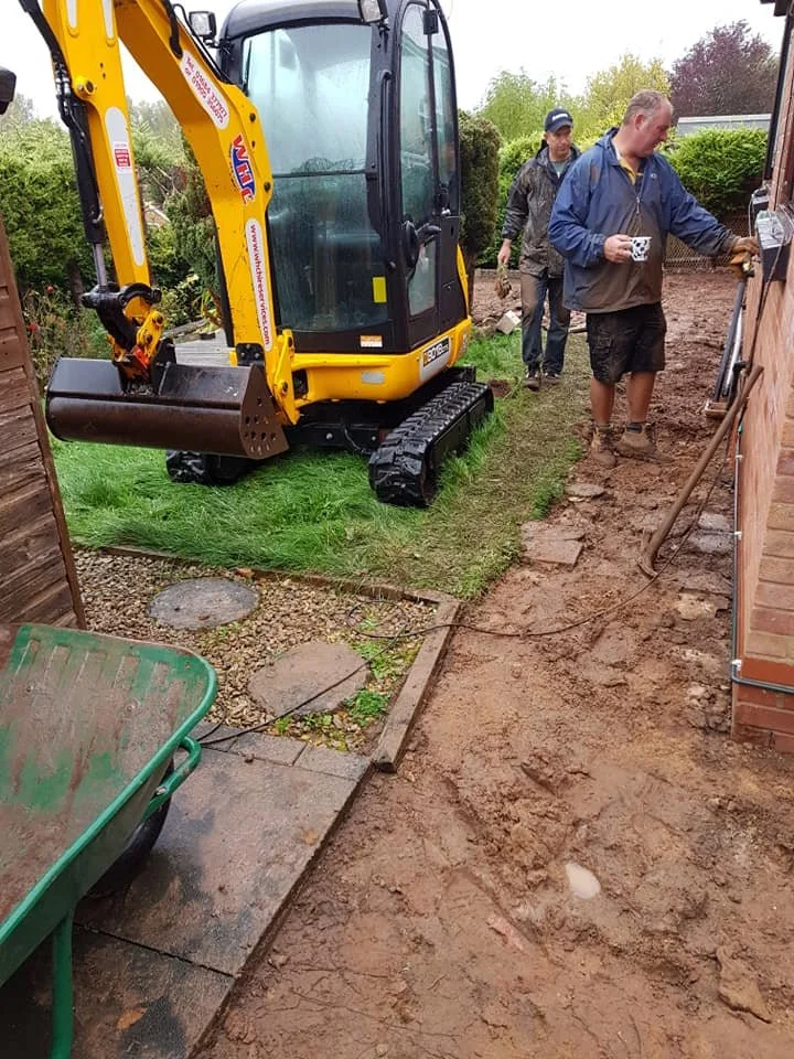 Two men working outside near a small yellow excavator next to a brick wall. One man is holding a mug and the other is walking behind. The ground appears to be dug up for a landscaping or construction project.