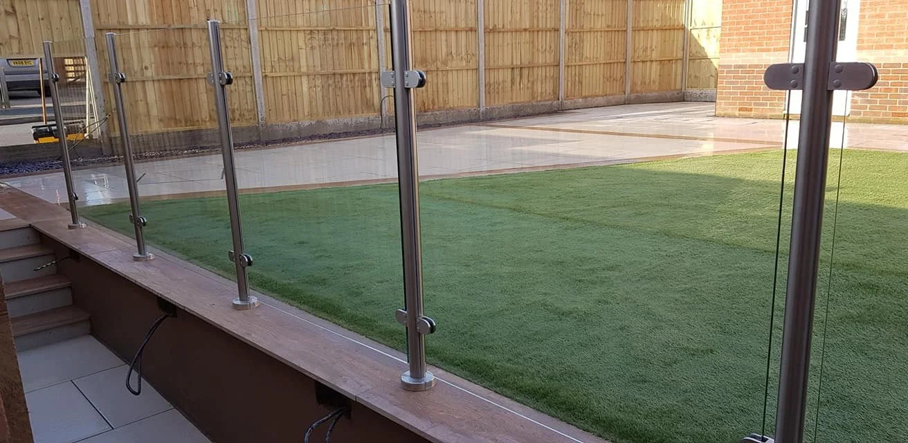 Backyard patio with artificial grass, wooden fencing, and a glass railing with metal posts.