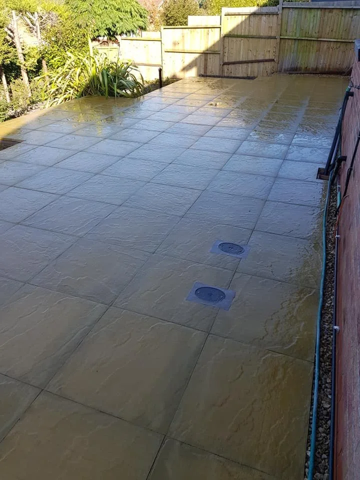 Wet outdoor tiled patio area with drain covers and a wooden fence in the background.