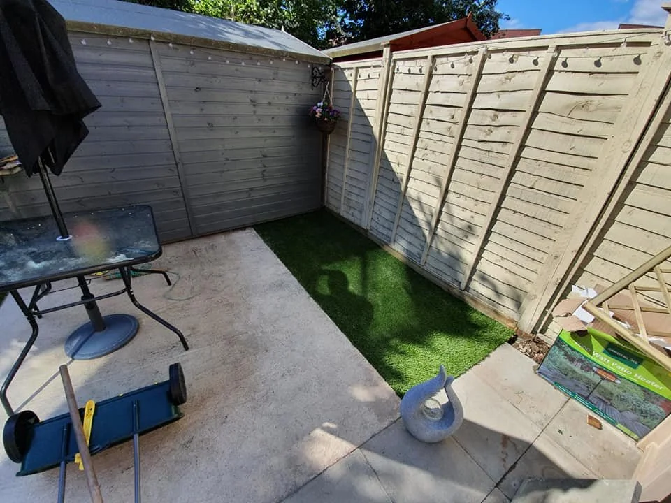 Small backyard patio area with a concrete floor, a patch of artificial grass, a table with an umbrella, a decorative swan statue, and a blocking fence.