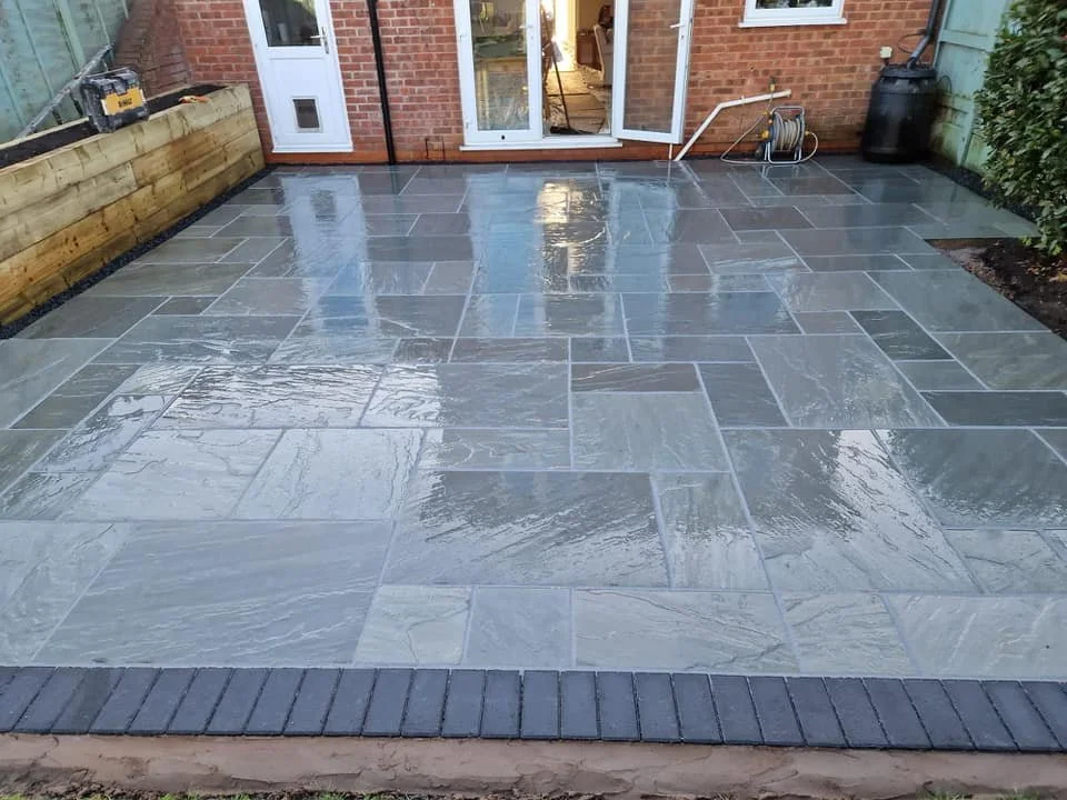 Recently installed gray stone patio in backyard, with wooden planter box on the left side, garden hose reel in the back right, and brick house with a sliding glass door.